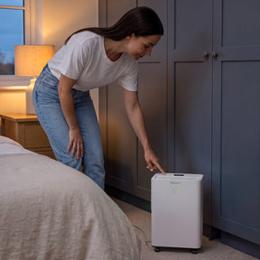 A woman in a white t-shirt and blue jeans points at a white portable dehumidifier on the floor in a bedroom setting.