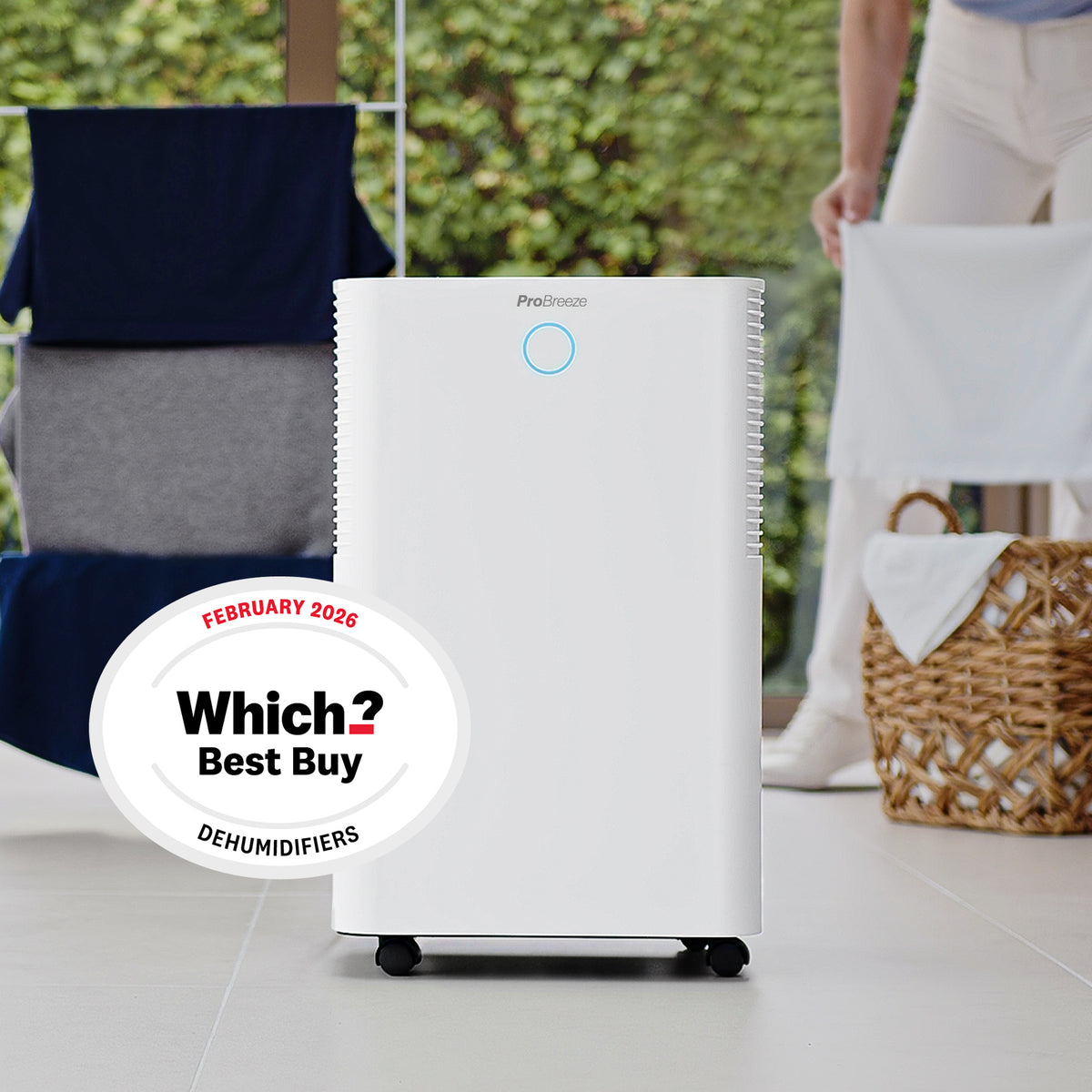 A white Predacons dehumidifier standing on a light tiled floor in a home setting, with a laundry basket, person, and chair blurred in the background. A 'Which? Best Buy February 2026' badge is overlaid on the left.