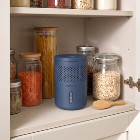 A blue Pro Breeze moisture absorber with a perforated front and a vertical level window sits on a kitchen shelf beside glass jars filled with pasta, lentils, and oats.