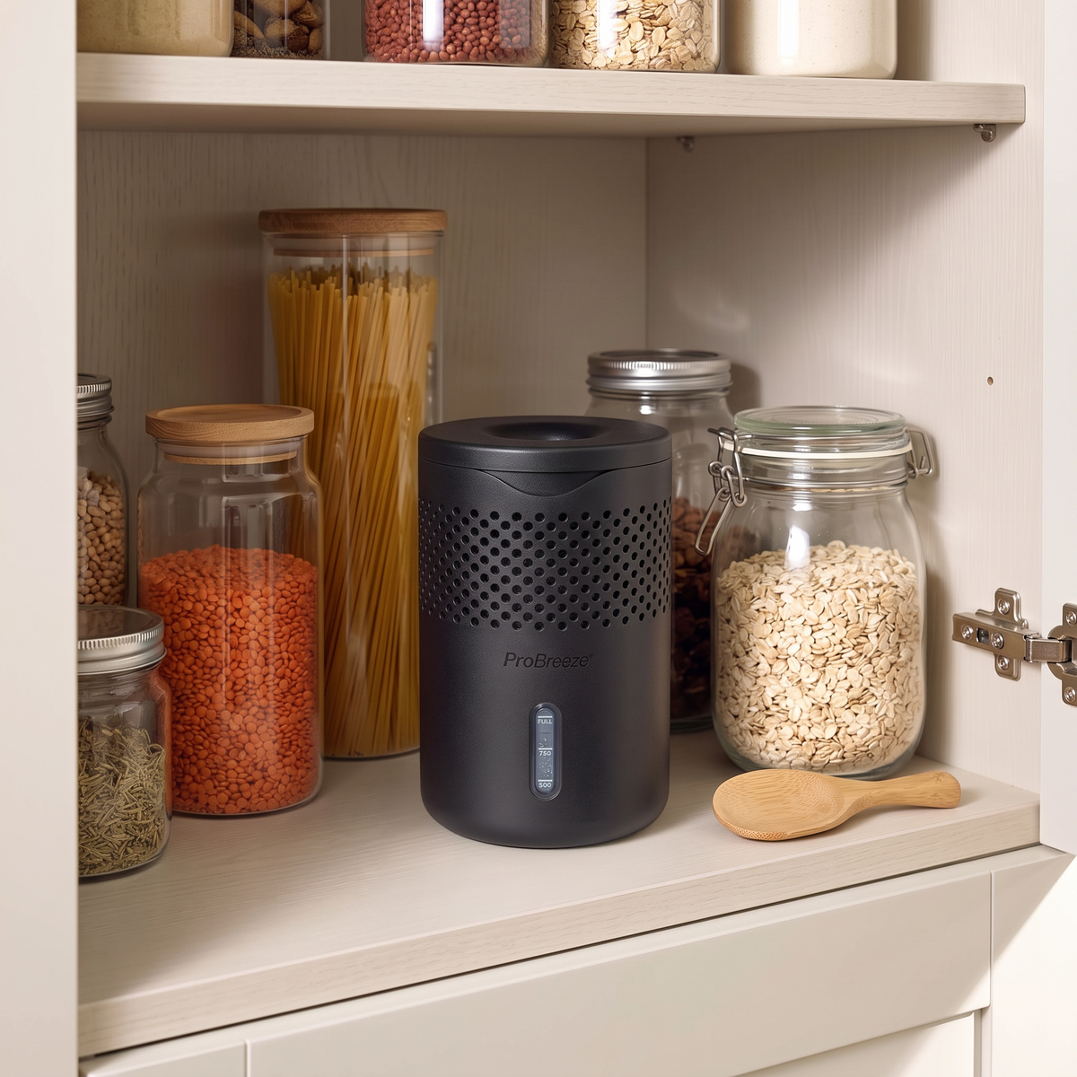 A black Pro Breeze moisture absorber sits on a kitchen cabinet shelf next to glass canisters filled with pasta, oats, and spices.
