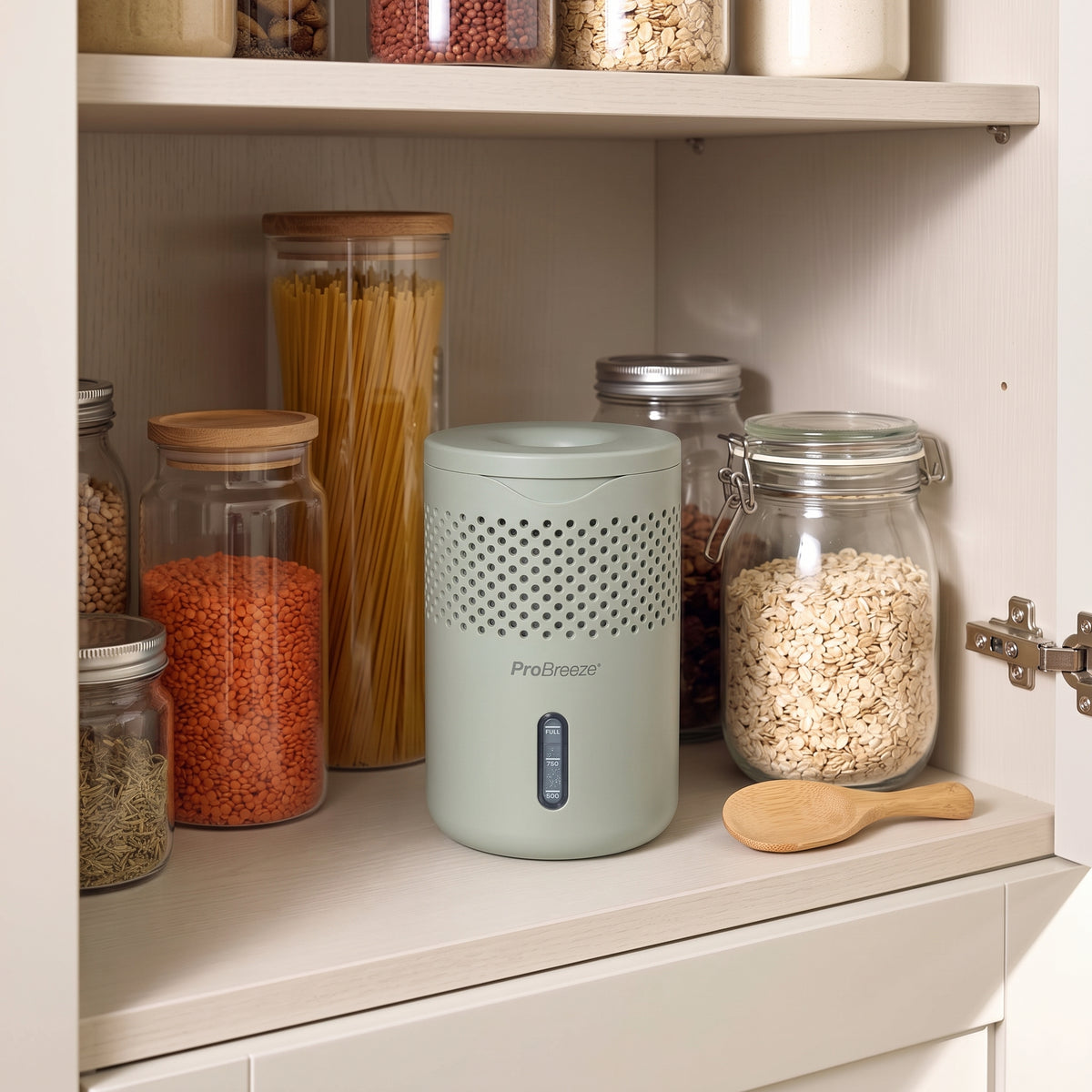 A light sage green PreBleezer air freshener dispenser sits on a kitchen shelf next to glass jars filled with pasta, lentils, and oats.