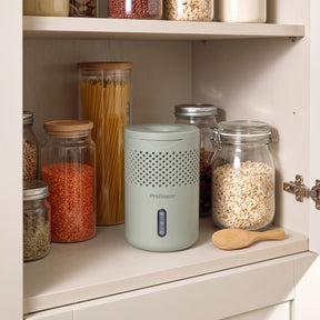 A light sage green PreBleezer air freshener dispenser sits on a kitchen shelf next to glass jars filled with pasta, lentils, and oats.