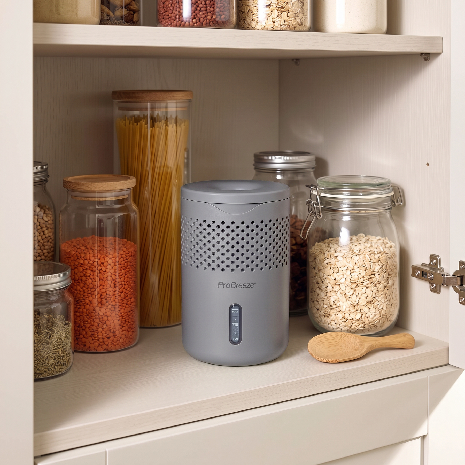 A gray Pro Breeze countertop moisture absorber with a perforated front and a vertical blue indicator window, shown on a kitchen shelf next to glass canisters of pasta, oats, and red beans.