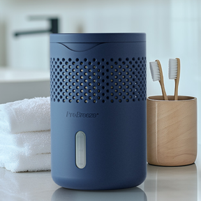 A blue cylindrical ProBreeze personal moisture absorber on a bathroom counter, flanked by a stack of white towels and a wooden toothbrush holder with two wooden toothbrushes.